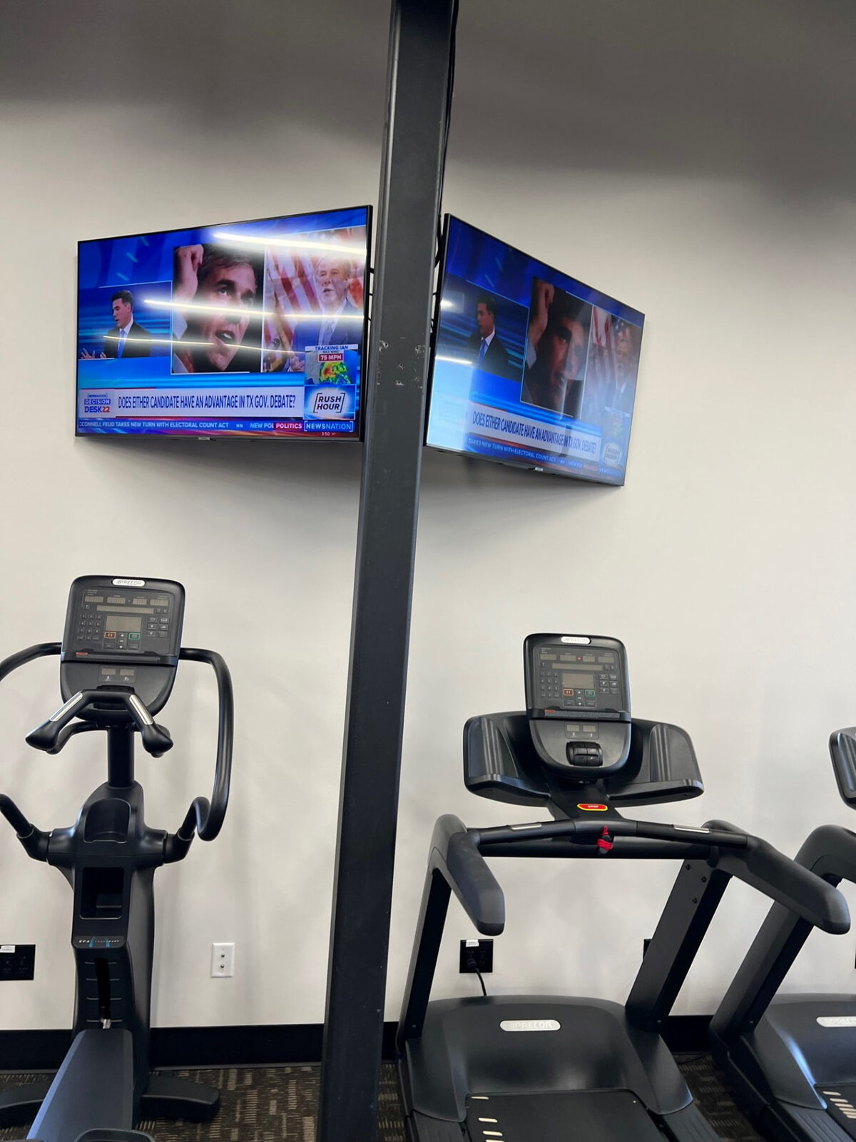 Paired televisions mounted on a pole in a fitness center cardio area